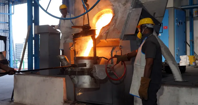 In a manufacturing facility, two employees melt metal and gather it in containers while wearing masks and safety gloves.